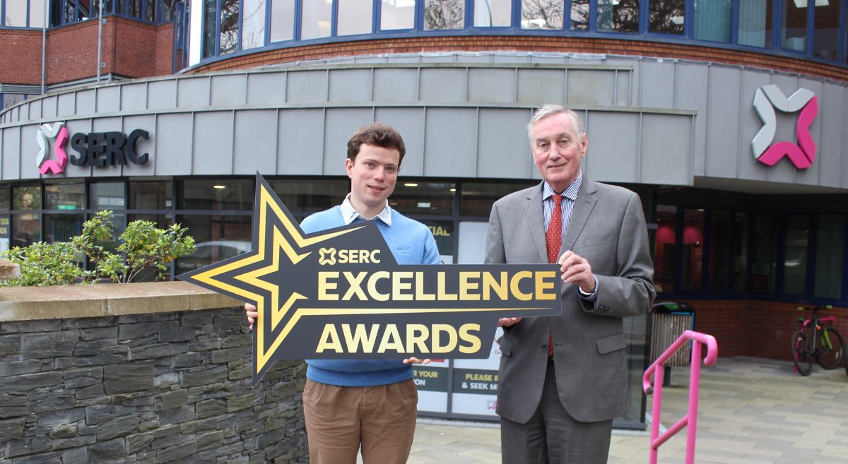 Picture of two males holding a star shaped sign which reads 'Excellence awards' with the Bangor campus in the background.